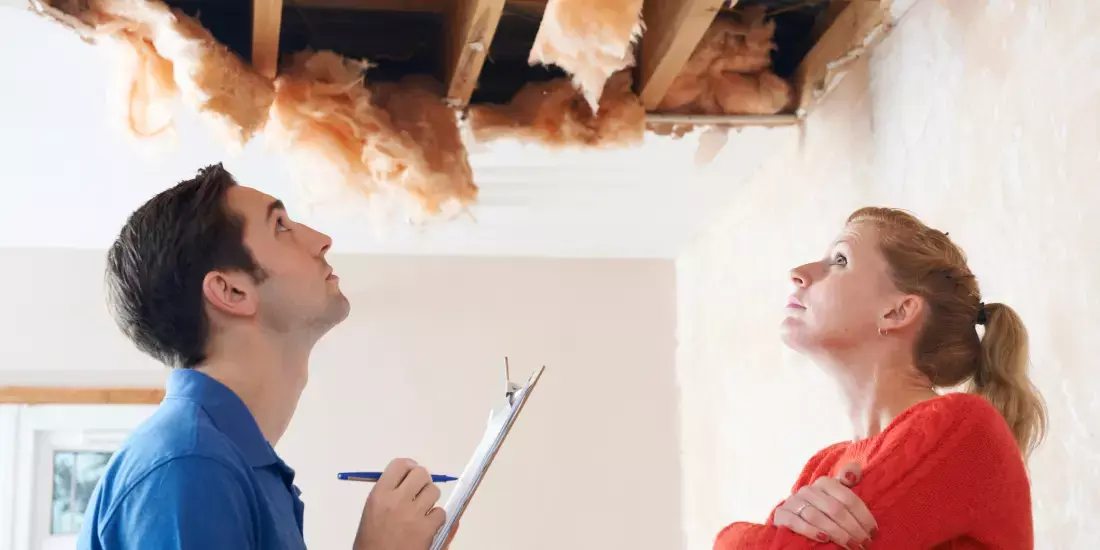 A man in blue with a clipboard and a woman in red assessing damage to the ceiling