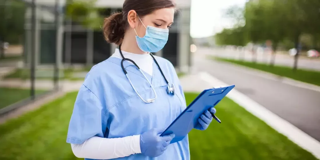 Female nurse stands in front of a hospital with a clipboard