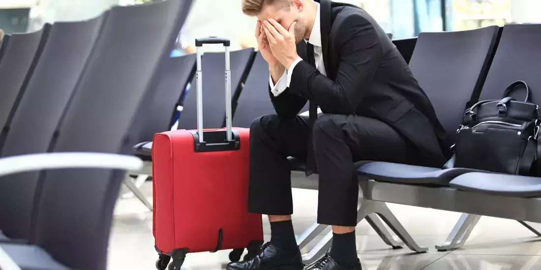 A man in a suit sits on a chair in an airport with his head in his hands. His luggage case is next to him and a black bag is on the adjacent chair