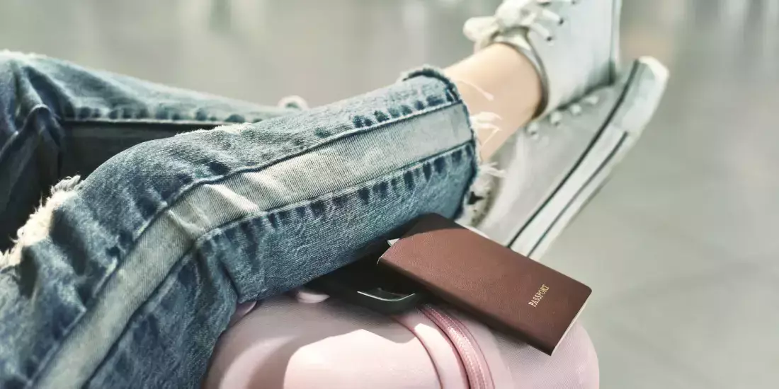 A woman in jeans with her legs resting on a pink luggage case and her passport on the side