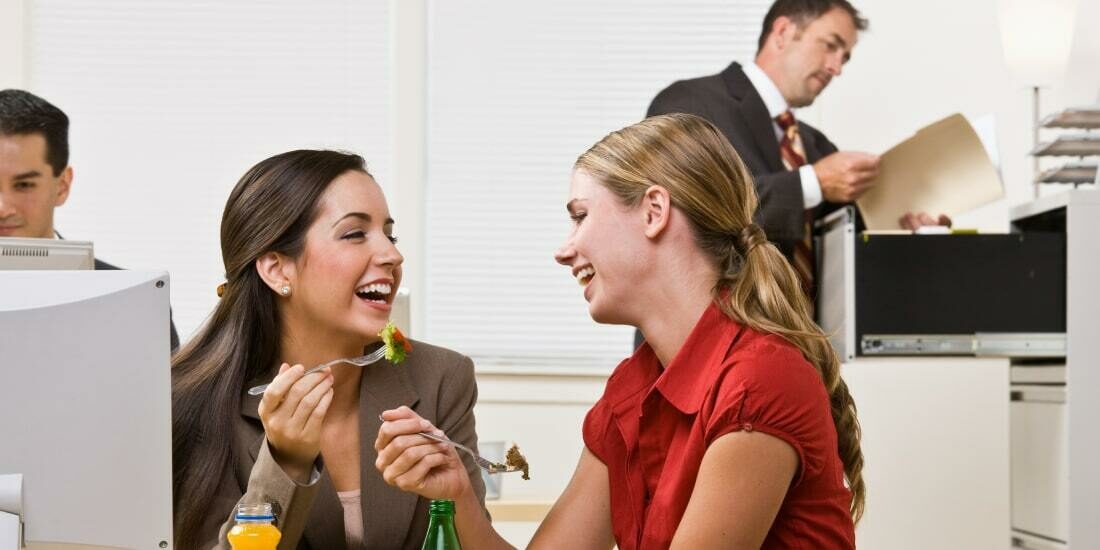 Two employees laugh and have lunch while two more employees work in the background